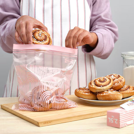 A person placing a cinnamon roll into an IKEA ISTAD resealable bag with a pale pink geometric pattern. The bag features a double-seal strip for secure closure. -80592710