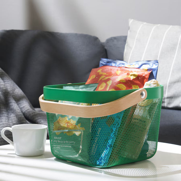 Dark green IKEA RISATORP basket, 25x26x18 cm (9 ¾x10 ¼x7 "), filled with assorted snacks and placed on a white surface. Features ventilated steel mesh, a sturdy wooden handle, and a stylish design for kitchen, office, or living room storage.-00588689