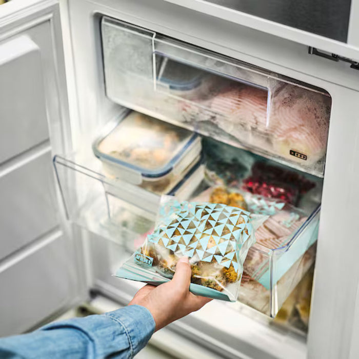 A person wearing a striped apron and a pink long-sleeve shirt places fresh vegetables into a pale grey-green IKEA ISTAD resealable bag. The bag has a geometric triangle pattern and a blue zip-seal.-50592716