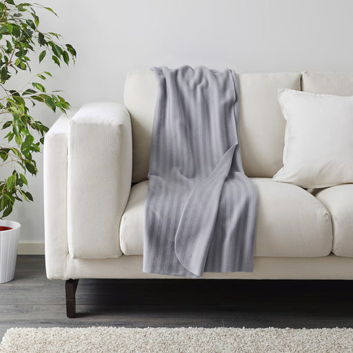 Cozy living room with a white sofa, draped with a light gray fleece blanket featuring subtle vertical stripes. A white pillow rests on the sofa, with a green potted plant nearby.