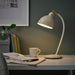 A stylish IKEA RÖDFLIK desk lamp in light beige with a vintage industrial design, featuring a shiny enamel-like finish, brass-colored accents, and an adjustable shade with a toggle switch for precise lighting control-20583921   