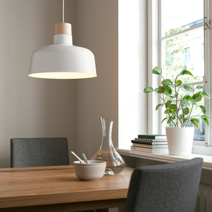 A bright and modern dining area featuring a wooden table with a glass carafe, a bowl with spoons, and two gray upholstered chairs. Above the table hangs a white pendant lamp with a wooden accent, blending minimalist design with natural elements. A window on the right lets in ample natural light, with a plant and books neatly placed on the windowsill, enhancing the cozy, stylish ambiance-90488398