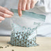 Person sealing an IKEA ISTAD resealable bag with pale grey-green triangle patterns. The bag contains seeds, highlighting its use for food storage.-50592716