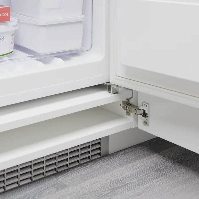 Close-up of the lower part of an open refrigerator door attached with a metal hinge. Inside the appliance are several white storage containers and an ice cream tub. The floor beneath is wooden, and the appliance features a vent at the bottom for air circulation.