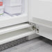 Close-up of the lower part of an open refrigerator door attached with a metal hinge. Inside the appliance are several white storage containers and an ice cream tub. The floor beneath is wooden, and the appliance features a vent at the bottom for air circulation.