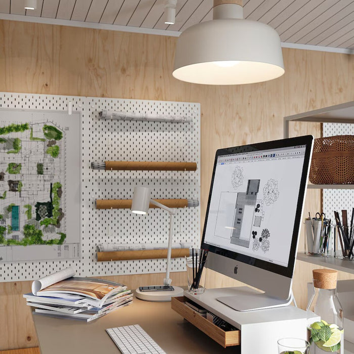A workspace featuring a large computer monitor displaying architectural or design software, including detailed building and landscaping plans. The desk is organized with a lamp, keyboard, and stacked books or magazines. A pegboard on the wall holds rolled-up plans and pinned landscape designs, creating a professional and creative environment-90488398