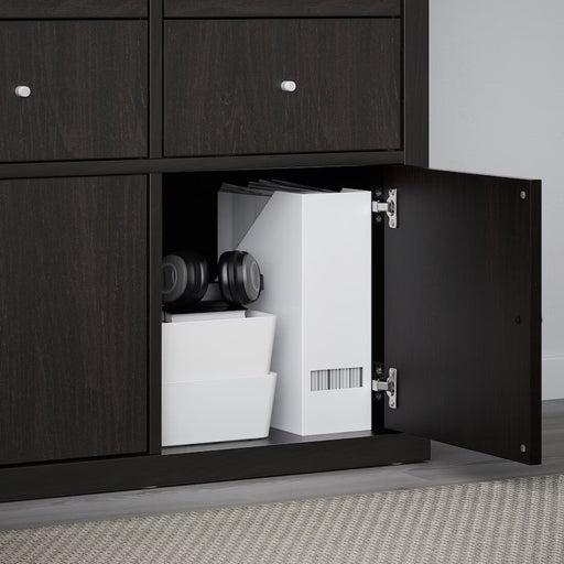 An open black-brown IKEA KALLAX insert with a door, revealing organized storage items inside. The compartment holds two white storage boxes stacked together, a pair of black headphones resting on top, and a white file organizer containing neatly arranged documents. The small, round knob on the door adds to its modern, minimalist design