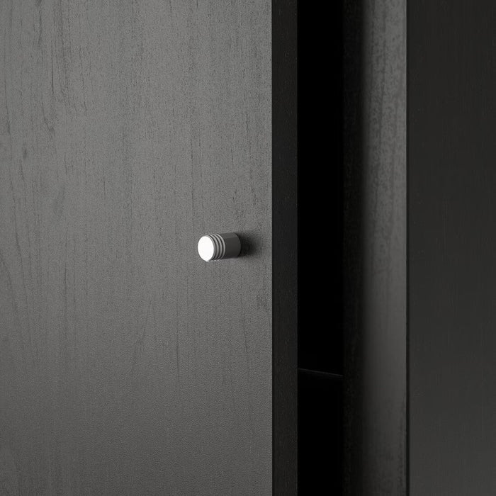 A close-up view of a black-brown IKEA KALLAX insert with a door, highlighting its smooth surface texture and small, round metallic knob centered on the door panel. The door is slightly ajar, revealing a dark interior, emphasizing the hidden storage functionality. The minimalist design complements modern home organization while providing a stylish solution for discreet storage.