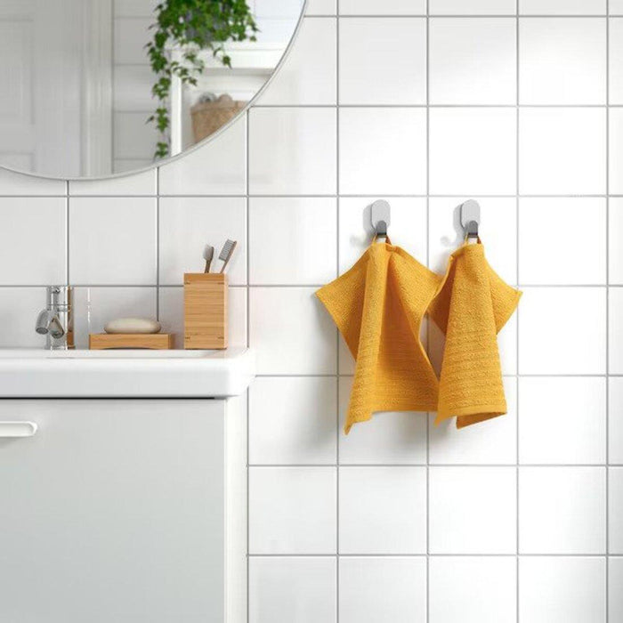 Bathroom scene with two golden-yellow IKEA VÅGSJÖN washcloths, 30x30 cm, hanging on hooks against a white tiled wall. The scene includes a white sink with a chrome faucet, a wooden holder with toothbrushes, and a soap dish with a bar of soap.90549518