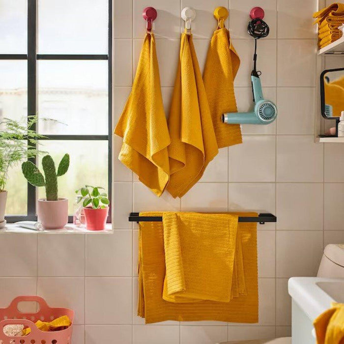 Bathroom setting with several golden-yellow towels and washcloths from IKEA's VÅGSJÖN collection. Three towels are hanging on hooks, and a washcloth is hanging on a towel rack below them.90549518