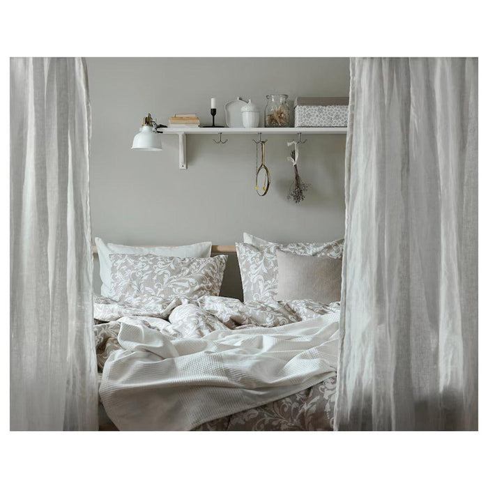 Cozy bedroom with a light-colored, patterned duvet and multiple pillows on the bed. Sheer curtains frame the bed, and a white shelf above holds books, a lamp, and decorative items. The overall aesthetic is neutral and inviting-00412614