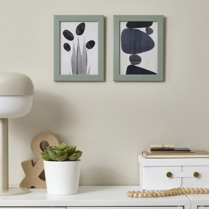 Two abstract art pieces in green frames on a wall above a white shelf with decor items.