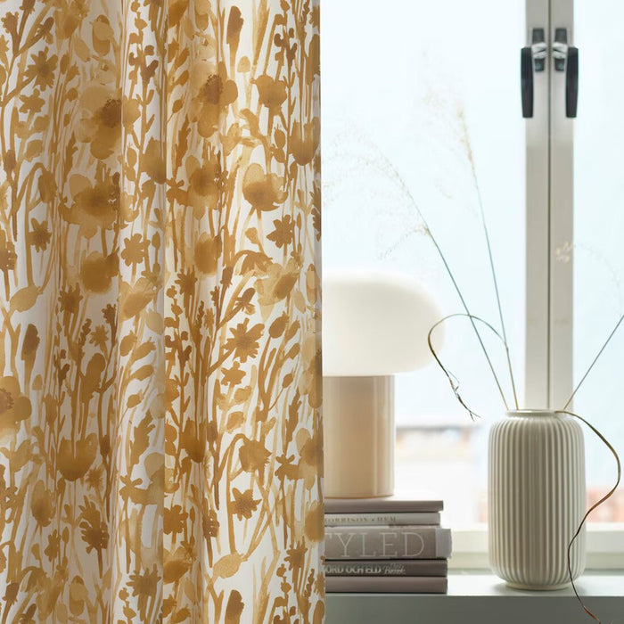 A pair of IKEA FLENTIMOTEJ curtains in light beige with a floral pattern, measuring 145x250 cm, are partially drawn across a window. The curtains feature large, stylized flowers and leaves in a slightly darker beige tone that creates a natural and soft aesthetic. Sunlight filters through the sheer fabric, casting a warm glow into the room. In the background, there’s a glimpse of an interior setting with a white table lamp on top of books and beside it, a textured white vase on the windowsill.