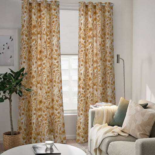  A pair of IKEA FLENTIMOTEJ curtains in light beige with a floral pattern, measuring 145x250 cm, are hanging in a well-lit room. The curtains are drawn open to let natural light in through the window they cover. The room is furnished with a modern aesthetic, featuring a neutral-colored sofa adorned with cushions, a small coffee table with a round tray on top, and a floor lamp. A potted plant adds greenery to the space. The wall behind the sofa has framed artwork that complements the room’s decor.