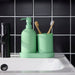 Green bathroom set with soap dispenser, toothbrush holder, and toothbrushes on a black tiled wall background.
