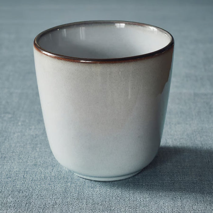A single IKEA GLADELIG espresso cup in light grey with a brown speckled rim, resting on a textured light blue fabric surface, with soft light falling on the cup."