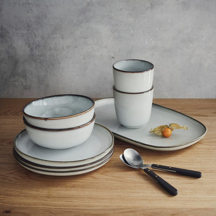 An IKEA GLADELIG stoneware dining set in light grey with brown rims, arranged on a light wooden table. The set includes stacked plates, bowls, two espresso cups, and a rectangular serving plate with a physalis fruit, next to two spoons