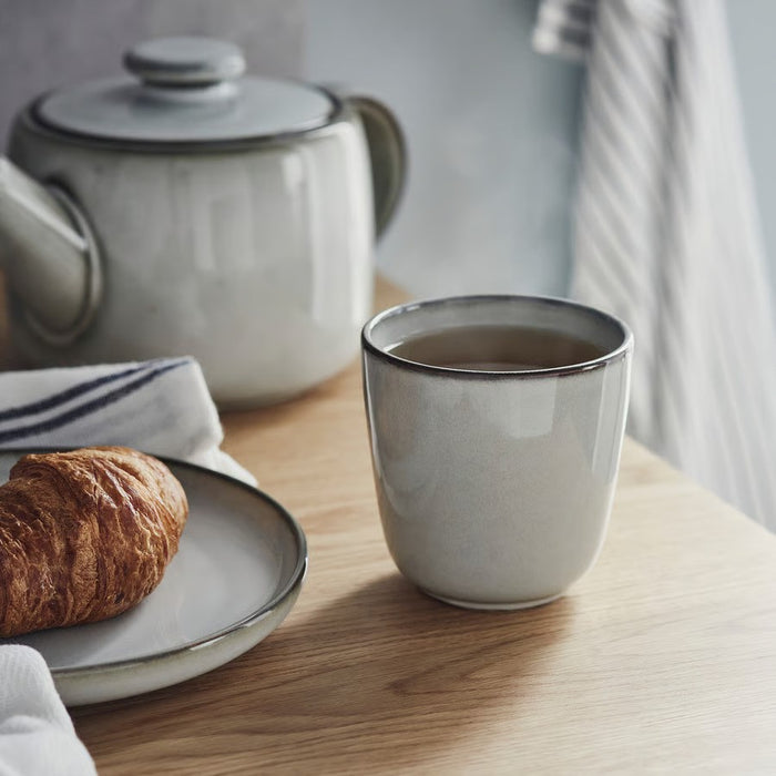 A grey IKEA GLADELIG espresso cup filled with a dark beverage sits on a light wooden table next to a plate with a golden-brown croissant and a folded white and blue striped towel. A matching grey ceramic teapot is blurred in the background.