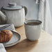 A grey IKEA GLADELIG espresso cup filled with a dark beverage sits on a light wooden table next to a plate with a golden-brown croissant and a folded white and blue striped towel. A matching grey ceramic teapot is blurred in the background.