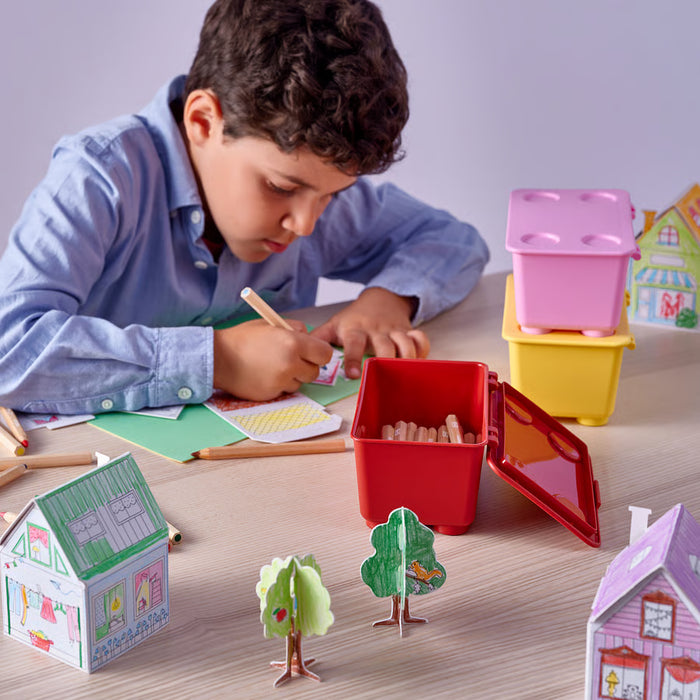Boy drawing at table with IKEA GLIS red yellow and pink boxes holding coloured pencils and craft supplies