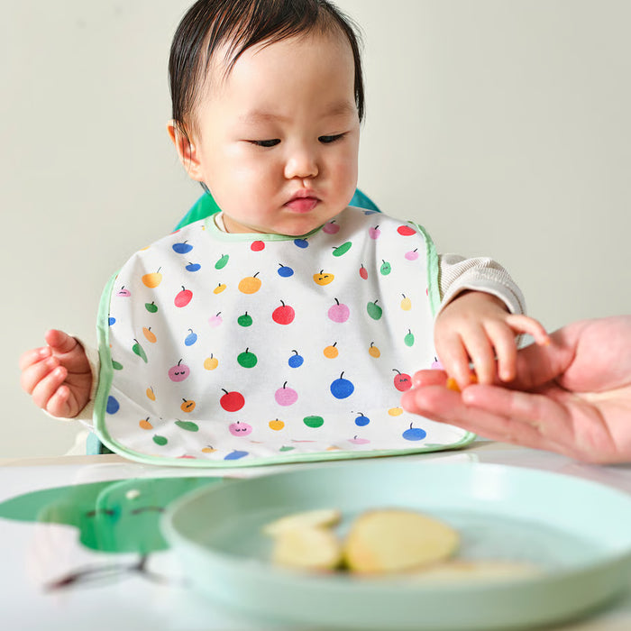 Baby at dining table wearing IKEA GRÖNFINK fruit-print bib being fed by adult caregiver