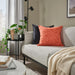 Living room with a gray sofa featuring orange and black patterned cushions, a small round table with plants, and a lamp.