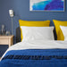 IKEA GURLI yellow cushion on a bed in a bedroom with blue walls and abstract wall art