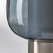 Close-up of the IKEA HÖGVIND table lamp, showing the smooth, translucent grey glass shade and the brushed nickel-plated base with a subtle horizontal texture."