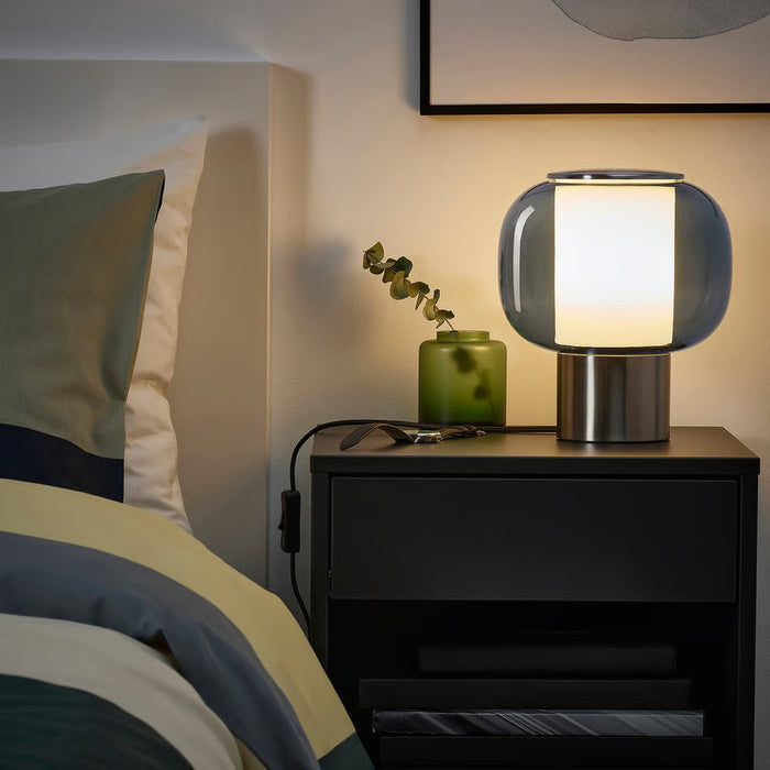 An illuminated IKEA HÖGVIND table lamp with a grey glass shade and nickel-plated base, sitting on a dark grey bedside table. Next to it are a small green vase with a plant, a charging cord, and books. A bed with green, yellow, and blue striped bedding is visible to the left."