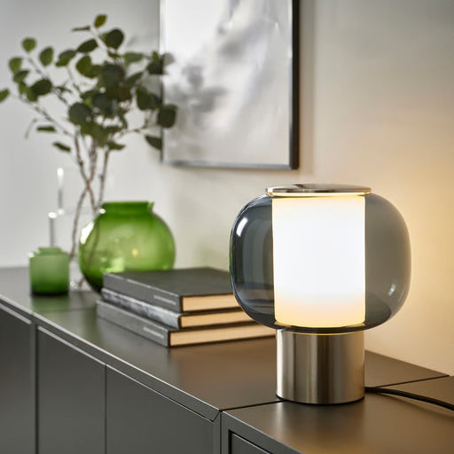 An illuminated IKEA HÖGVIND table lamp with a glowing white inner cylinder and a dark grey glass outer shade, resting on a dark grey sideboard next to a green vase with eucalyptus branches and a stack of books, in a dimly lit room.