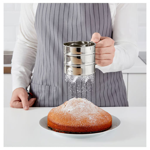  Person in grey apron dusting a white cake with icing sugar using IKEA IDEALISK flour sifter