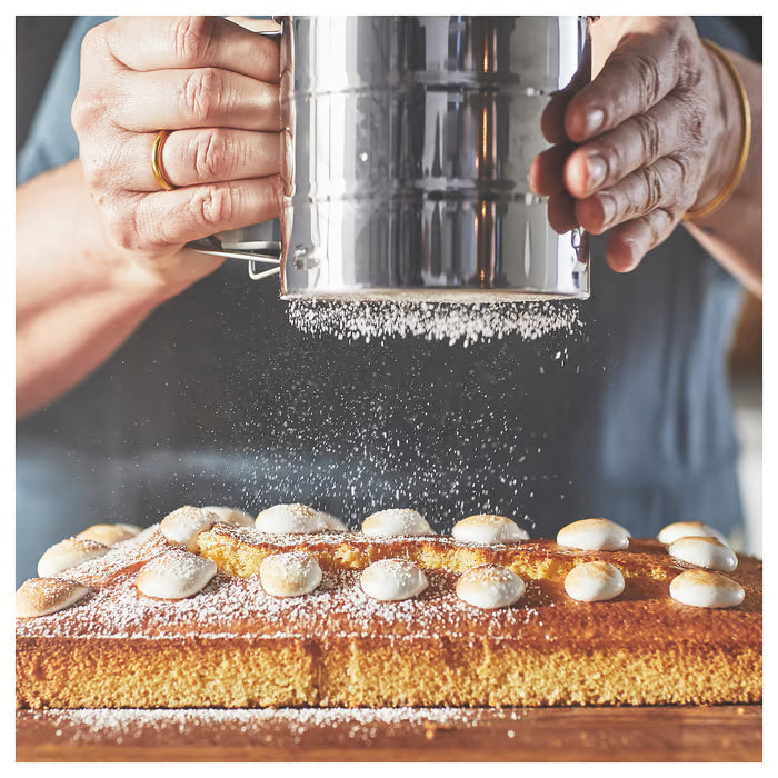 Golden sliced cake dusted with sugar alongside baking tools, featuring IKEA IDEALISK flour sifter