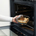 Person using oven gloves to place IKEA 365+ glass food container into oven, showing oven-safe use