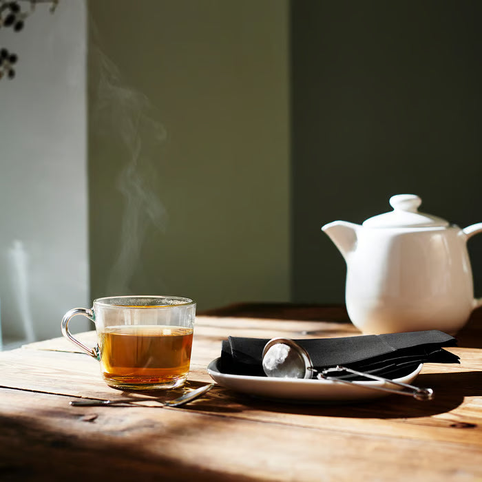 IKEA 365+ glass mug filled with steaming tea on wooden table next to white teapot and silver spoon
