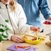 Child holding carrot beside open IKEA KAPTENSFISK yellow lunch box with sandwich and veggies on wooden table
