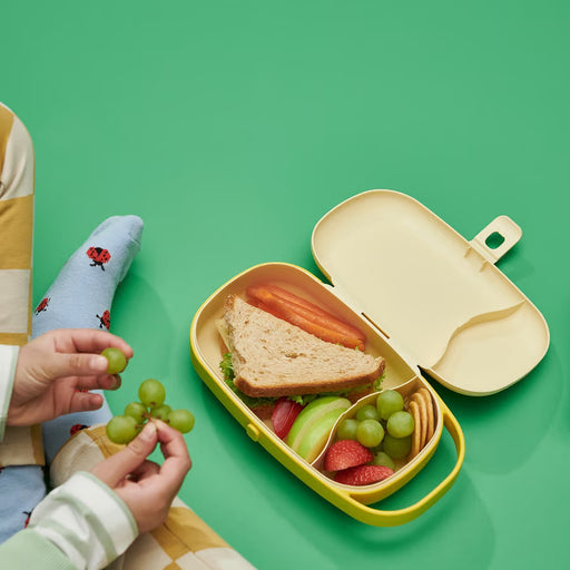 Child's hand holding IKEA KAPTENSFISK yellow lunch box with sandwich, fruit and crackers in separate compartments