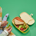 Child's hand holding IKEA KAPTENSFISK yellow lunch box with sandwich, fruit and crackers in separate compartments
