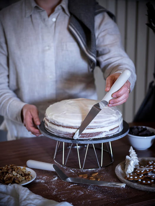 Person using IKEA LÄTTBAKAD offset spatula to cut a white frosted cake on a wire stand