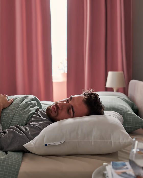 Person sleeping on IKEA LAPPTÅTEL white pillow side view red curtains bedside lamp on