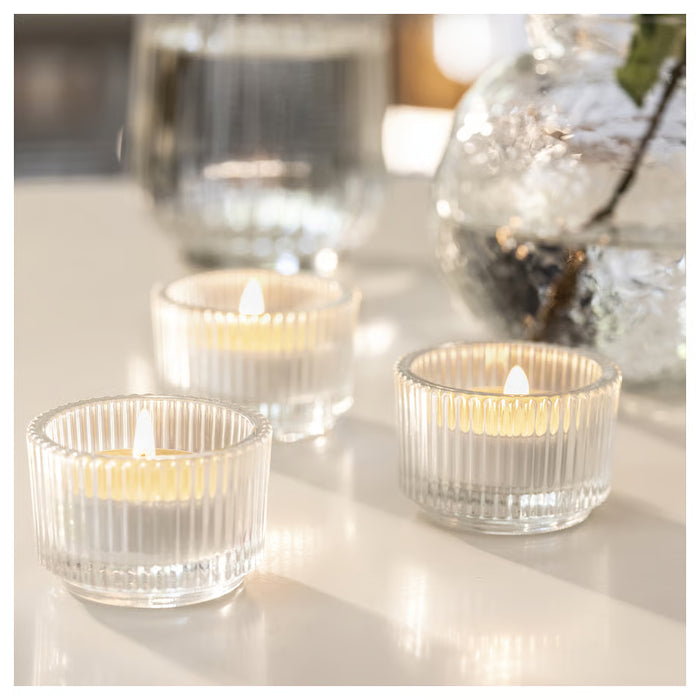 Three lit LÖVSKOGSLUND glass tealights on white surface with blurred vase in background