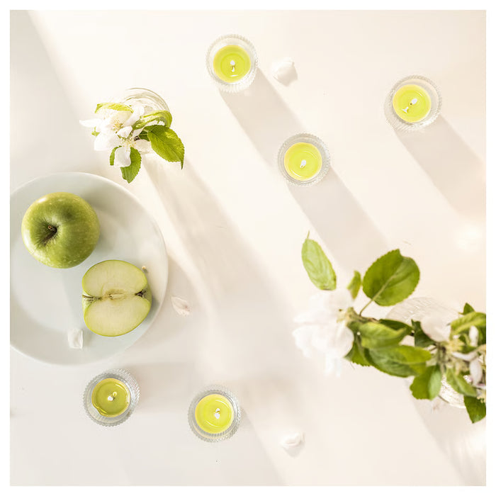 Six lit LÖVSKOGSLUND green apple scented tealights arranged with apple slices on bright table