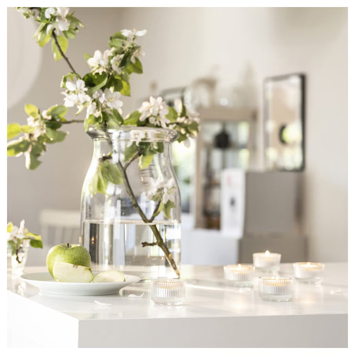 Lit LÖVSKOGSLUND tealights on white counter with glass vase, blooming branches and green apples