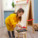 Young girl crafting with IKEA MÅLA art set including brushes, scissors, and markers on a wooden stool