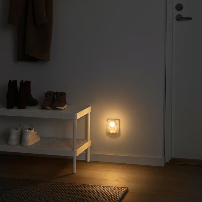 IKEA MÖRKRÄDD white LED nightlight plugged into a wall socket in a dark hallway, emitting a soft warm glow