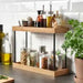 IKEA NÅLBLECKA two-tier kitchen countertop organiser made of bamboo and black metal, filled with spice jars, oil bottles, and condiments, on a grey kitchen counter with white subway tile backsplash-90564617