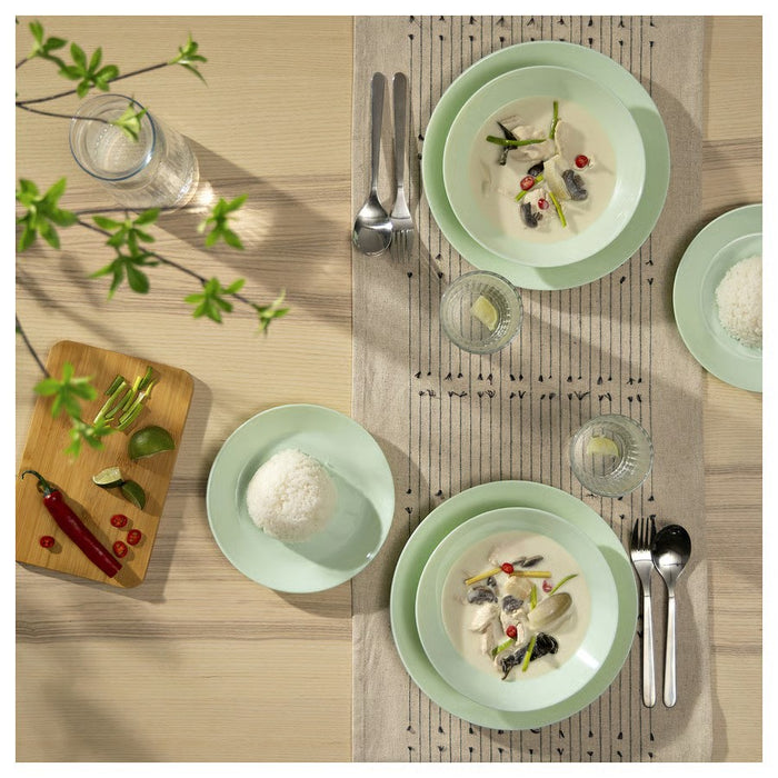 A breakfast setup on a wooden table featuring a green bowl, green plates with a croissant and a slice of cheese, a glass jar of granola, a pitcher of milk, sliced bread on a wooden cutting board, a fork, and a striped cloth napkin, illuminated by natural light for a warm, inviting feel."