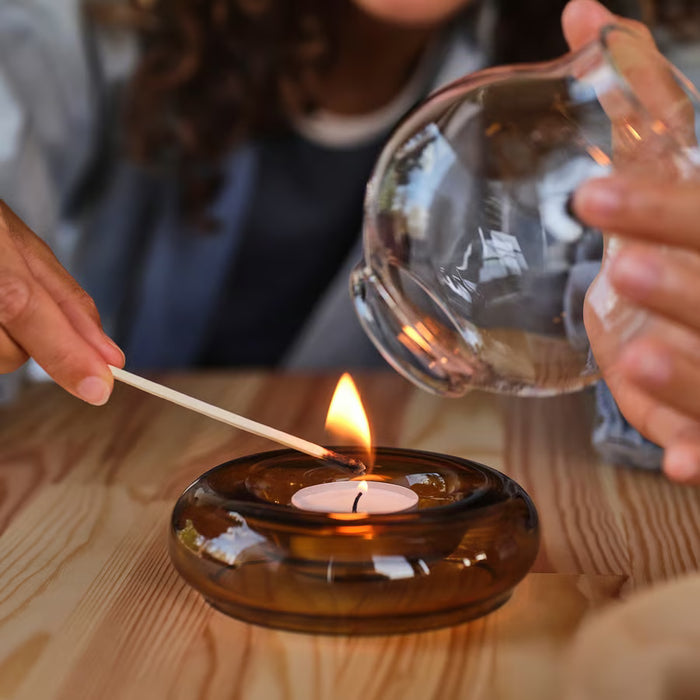 Person lighting a tealight in IKEA OMMJÄNGE brown glass holder on wooden table warm glowing flame