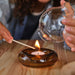 Person lighting a tealight in IKEA OMMJÄNGE brown glass holder on wooden table warm glowing flame
