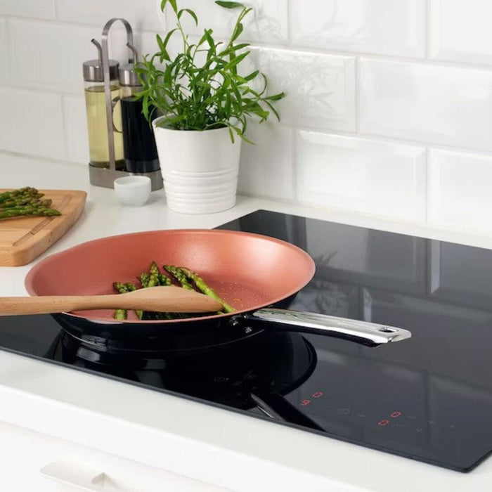 Copper frying pan with asparagus on a black induction cooktop in a kitchen.