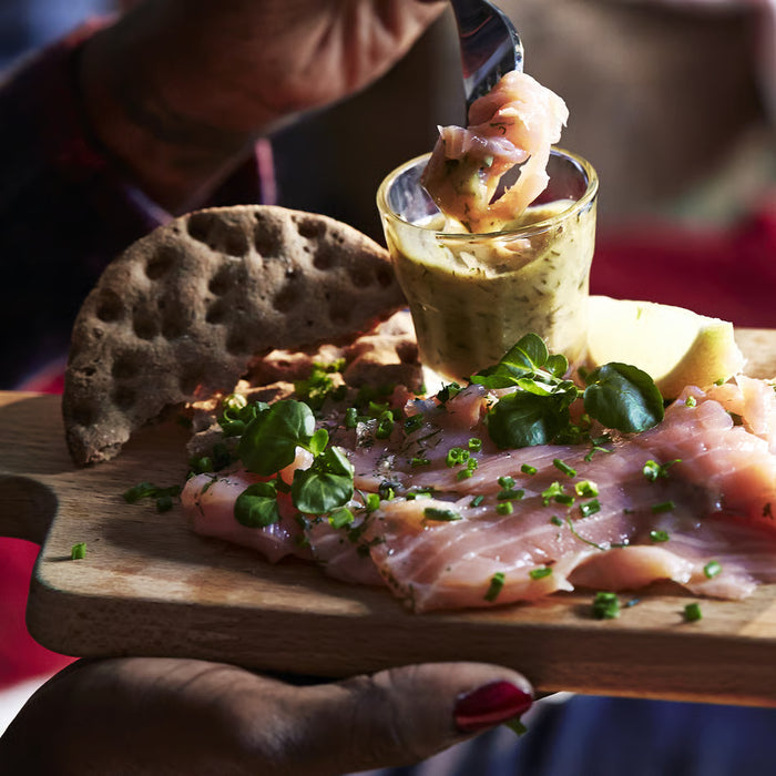 Person dipping bread into sauce using IKEA POKAL snaps glass alongside smoked fish and greens on wooden board